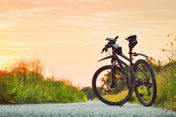 Naklejka na ściany i meble The mountain bike stands on a gravel bike path among green vegetation illuminated by the rays of the setting sun.