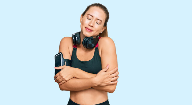 Beautiful blonde woman wearing gym clothes and using headphones hugging oneself happy and positive, smiling confident. self love and self care