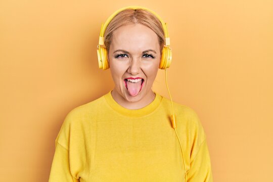 Young blonde woman listening to music using headphones sticking tongue out happy with funny expression. emotion concept.