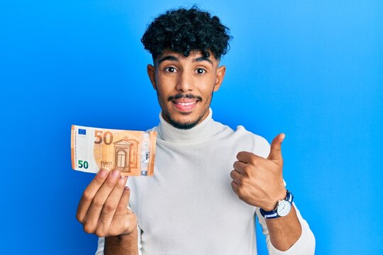 Young arab handsome man holding 50 euro banknote smiling happy and positive, thumb up doing excellent and approval sign