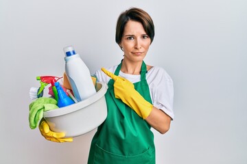 Young brunette woman with short hair wearing apron holding cleaning products pointing aside worried and nervous with forefinger, concerned and surprised expression