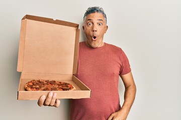 Handsome middle age mature man holding delivery pizza box scared and amazed with open mouth for...