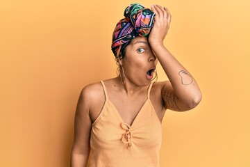 Young hispanic girl wearing hair turban over yellow background surprised with hand on head for...