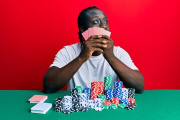 Handsome young black man playing gambling poker covering face with cards smiling looking to the side and staring away thinking.