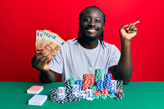 Handsome young black man playing poker holding 50 euros banknotes smiling happy pointing with hand and finger to the side