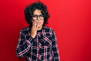 Fototapeta na wymiar Young hispanic woman with curly hair wearing casual clothes and glasses touching mouth with hand with painful expression because of toothache or dental illness on teeth. dentist