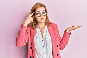 Young caucasian woman wearing business style and glasses confused and annoyed with open palm showing copy space and pointing finger to forehead. think about it.