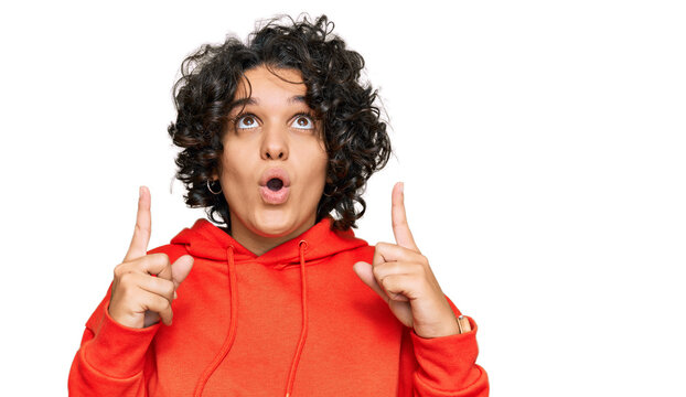 Young hispanic woman with curly hair wearing casual sweatshirt amazed and surprised looking up and pointing with fingers and raised arms.