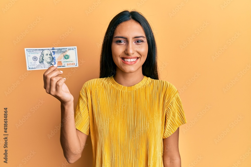 Wall mural Young brunette woman holding 100 dollars banknote looking positive and happy standing and smiling with a confident smile showing teeth