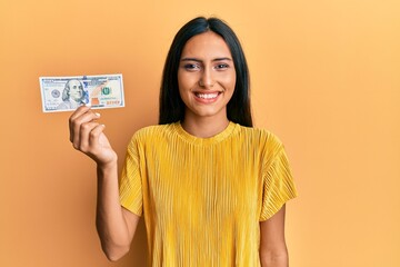 Young brunette woman holding 100 dollars banknote looking positive and happy standing and smiling with a confident smile showing teeth