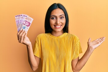 Young brunette woman holding 50 mexican pesos banknotes celebrating achievement with happy smile...