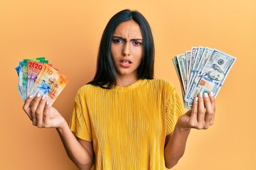 Young brunette woman holding bunch of dollars and swiss francs banknotes in shock face, looking skeptical and sarcastic, surprised with open mouth