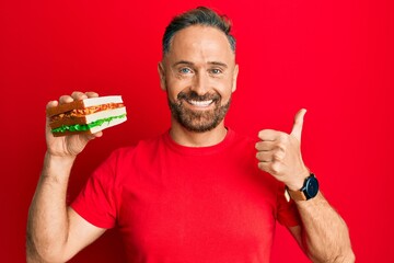 Handsome middle age man eating sandwich smiling happy and positive, thumb up doing excellent and approval sign