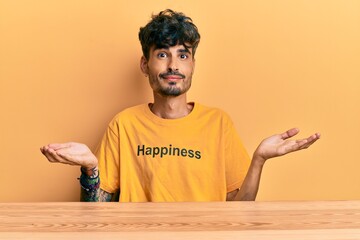 Young hispanic man wearing tshirt with happiness word message sitting on the table clueless and...