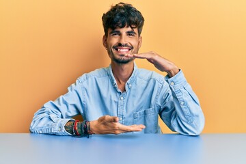 Young hispanic man wearing casual clothes sitting on the table gesturing with hands showing big and large size sign, measure symbol. smiling looking at the camera. measuring concept.