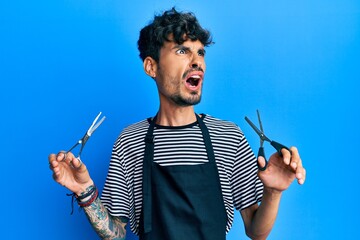 Young hispanic man wearing barber apron holding scissors angry and mad screaming frustrated and...