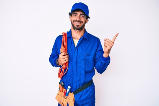 Handsome young man with curly hair and bear wearing builder jumpsuit uniform and electric cables smiling happy pointing with hand and finger to the side