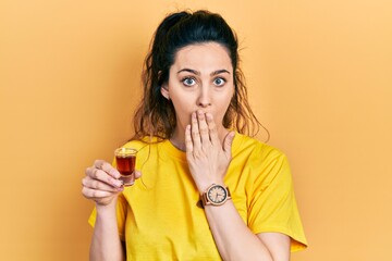 Young hispanic woman drinking whiskey shot covering mouth with hand, shocked and afraid for...