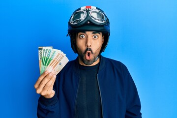 Young hispanic man wearing motorcycle helmet holding south korea won scared and amazed with open...