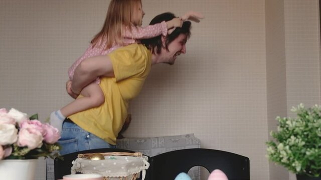 Easter, family, holiday and children concept. Dad in bunny ears plays like easter bunny, jumping with daughter. Happy family preparing for Easter