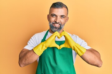 Middle age handsome man wearing cleaner apron and gloves smiling in love doing heart symbol shape...