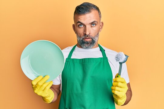 Middle age handsome man wearing apron holding scourer washing dishes in shock face, looking skeptical and sarcastic, surprised with open mouth