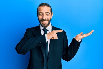 Attractive man with long hair and beard wearing business suit and tie amazed and smiling to the camera while presenting with hand and pointing with finger.