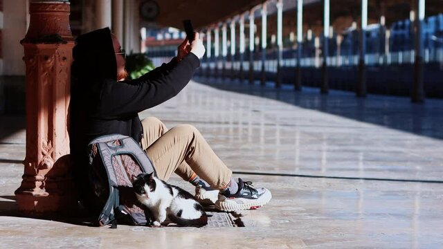 Young man traveller sitting on the station with street cats - using his phone and taking pictures of cats