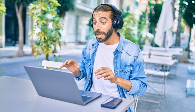 Young middle eastern man doing video call using laptop and headphones at coffee shop terrace.
