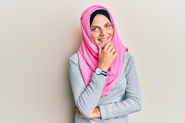 Young caucasian woman wearing traditional islamic hijab scarf looking confident at the camera smiling with crossed arms and hand raised on chin. thinking positive.