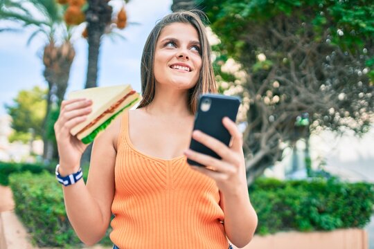 Young middle east girl smiling happy using smartphone and eating sandwich at the city.