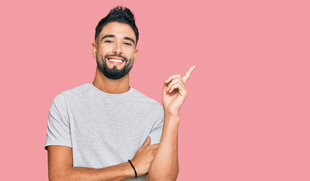 Young man with beard wearing casual grey tshirt with a big smile on face, pointing with hand and finger to the side looking at the camera.