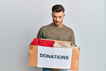 Young redhead man volunteer holding donations box skeptic and nervous, frowning upset because of...