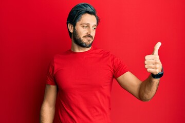 Fototapeta premium Young hispanic man wearing casual red t shirt looking proud, smiling doing thumbs up gesture to the side