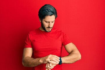 Young hispanic man wearing casual red t shirt checking the time on wrist watch, relaxed and confident