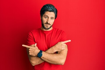 Young hispanic man wearing casual red t shirt pointing to both sides with fingers, different direction disagree