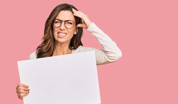 Young brunette woman holding blank empty banner stressed and frustrated with hand on head, surprised and angry face