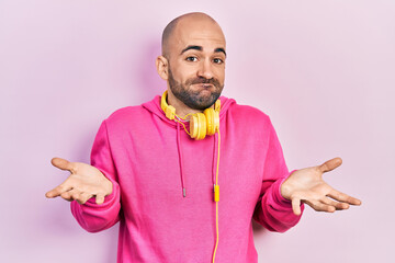 Young bald man wearing gym clothes and using headphones clueless and confused expression with arms...