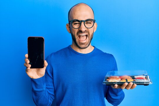 Young hispanic man holding take away food showing smartphone screen smiling and laughing hard out loud because funny crazy joke.