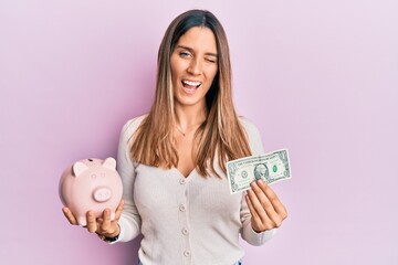 Brunette young woman holding one dollar banknote and piggy bank winking looking at the camera with...