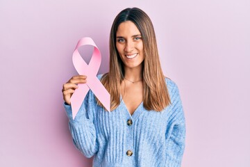 Brunette young woman holding pink cancer ribbon looking positive and happy standing and smiling with a confident smile showing teeth