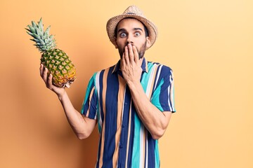 Young handsome man with beard wearing summer hat and shirt holding pineapple covering mouth with...