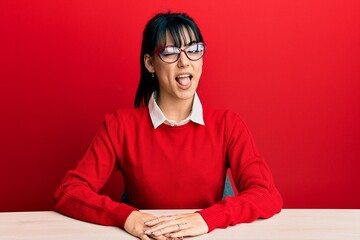 Young brunette woman with bangs wearing glasses sitting on the table winking looking at the camera...