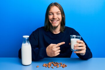 Handsome caucasian man with long hair drinking healthy almond milk smiling happy pointing with hand...