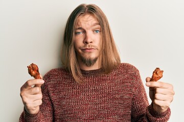 Handsome caucasian man with long hair eating chicken wings skeptic and nervous, frowning upset...