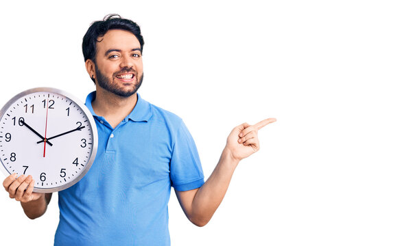 Young hispanic man holding big clock smiling happy pointing with hand and finger to the side