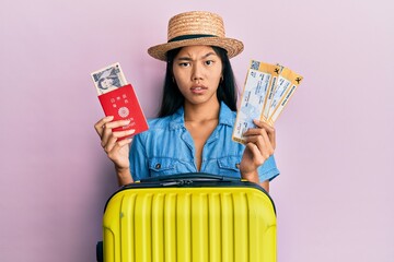 Young chinese woman holding boarding pass and passport in shock face, looking skeptical and...