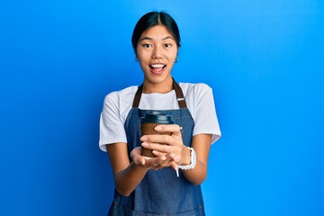 Young chinese woman wearing waiter apron holding cup of coffee celebrating crazy and amazed for success with open eyes screaming excited.