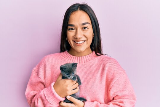 Young hispanic girl smiling happy holding cute cat over isolated pink background.