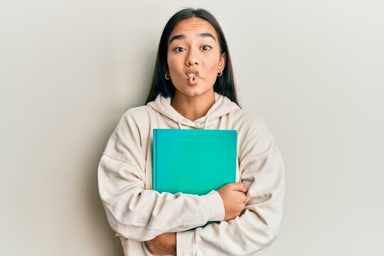 Young asian woman holding book making fish face with mouth and squinting eyes, crazy and comical.